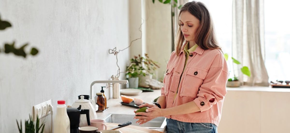 Young woman in pink jacket preparing avocado toast in a sunlit kitchen with houseplants.
