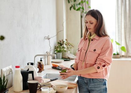 Young woman in pink jacket preparing avocado toast in a sunlit kitchen with houseplants.